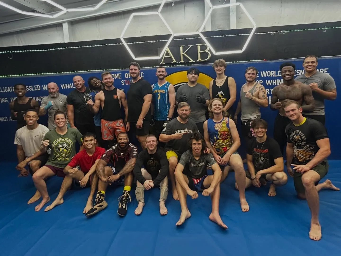 About two dozen Elite Combat Academy members on blue mats under hexagon lights and an AKB sign
